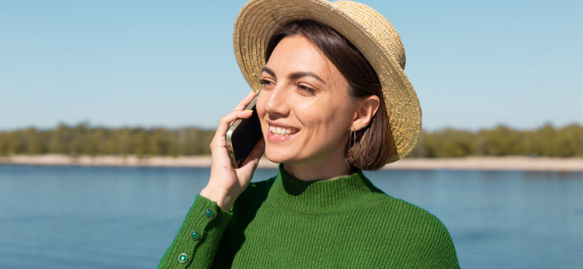 Stylish woman in green casual sweater and hat outdoor on bridge with river view at warm sunny summer day talks on mobile phone smile and laugh enjoy conversation