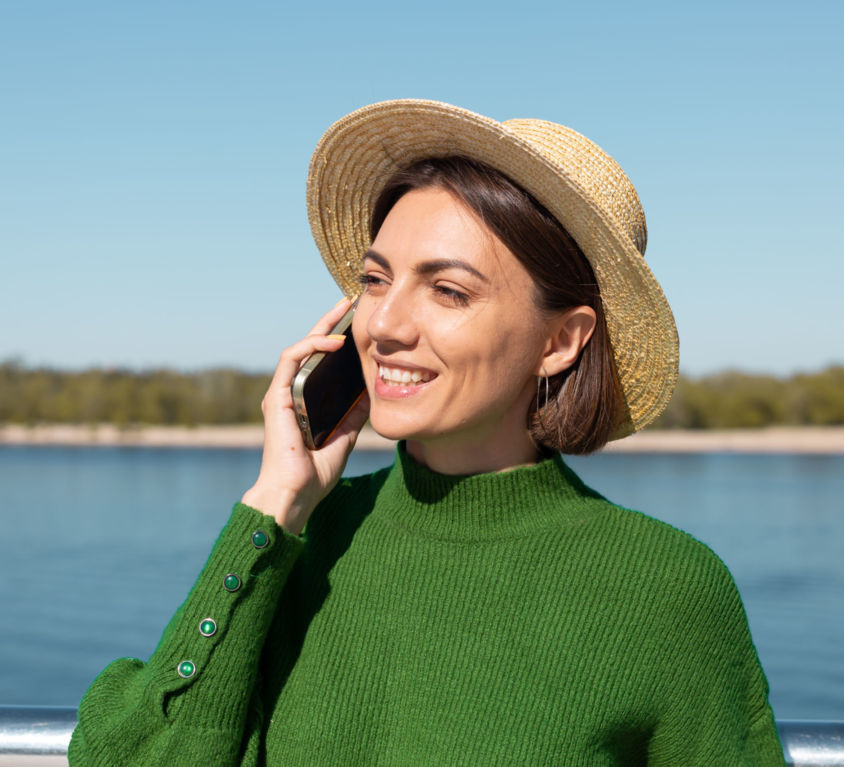 Stylish woman in green casual sweater and hat outdoor on bridge with river view at warm sunny summer day talks on mobile phone smile and laugh enjoy conversation